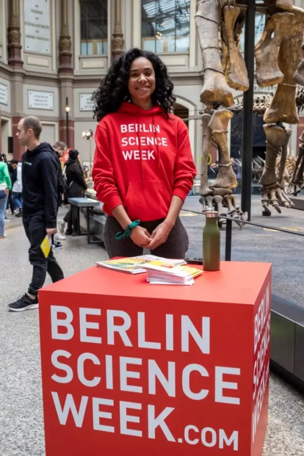 Foto: Volunteer mit schwarzen Locken und roten Hoodie vor Würfel mit "Berlin Science Week" Aufdruck